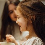 A little girl sitting in front of a sink next to a woman